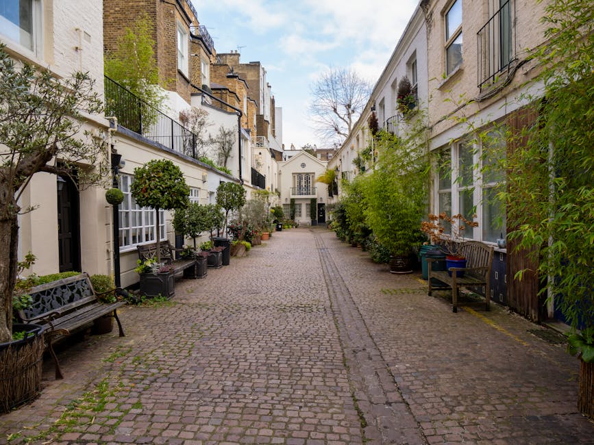 A cobbled alleyway in a residential area with white terraced houses on both sides. The pavement is lined with potted trees and plants, some in black containers and others in decorative planters, creating a green border along the buildings. There are several wooden benches along the alley, one on each side, and a few windows with black railings visible. The alley leads to a small white building with a window and door at the far end, under a partly cloudy sky. The environment is calm and well-maintained, suitable for house removals or furniture transport, with natural lighting highlighting the details of the outdoor setting. This scene relates to the process of home relocation and packing, as Movers West Kensington provide professional removals services through narrow access routes like this residential alley.