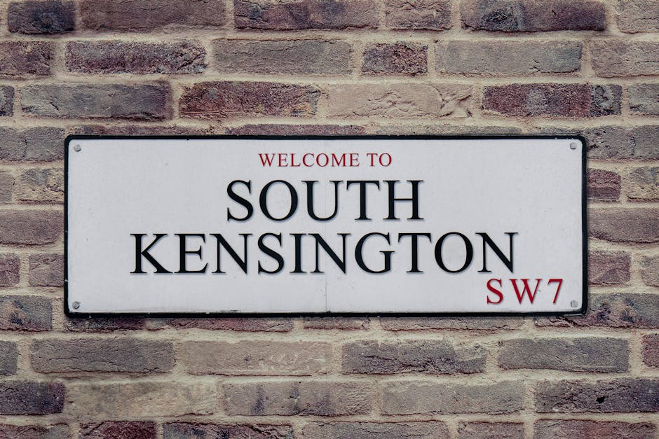 The image displays a rectangular white metal street sign mounted on a light brown brick wall, featuring black uppercase text that reads 'WELCOME TO SOUTH KENSINGTON' with the postal code 'SW7' in red at the bottom right corner. The sign is attached with four screws, one at each corner. The surrounding brick wall has a traditional pattern with a mix of darker and lighter shades, providing a textured background. This type of signage is typical for identifying neighbourhoods in London and is relevant to home relocation or moving services in the South Kensington area, under the domain of Movers West Kensington. The lighting appears natural, highlighting the details of the brickwork and the sign, which may be part of the initial or final stages of a house removal process in the Kensington vicinity.