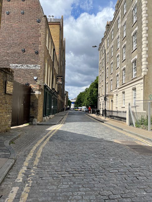 A narrow cobbled street in Notting Hill Gate, WEST KENSINGTON, lined with closely packed terraced houses featuring white, cream, and brick facades. Some houses have small balconies with black railings